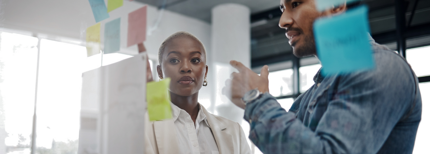 A professional woman and man standing together, collaborating and reviewing sticky notes on a board